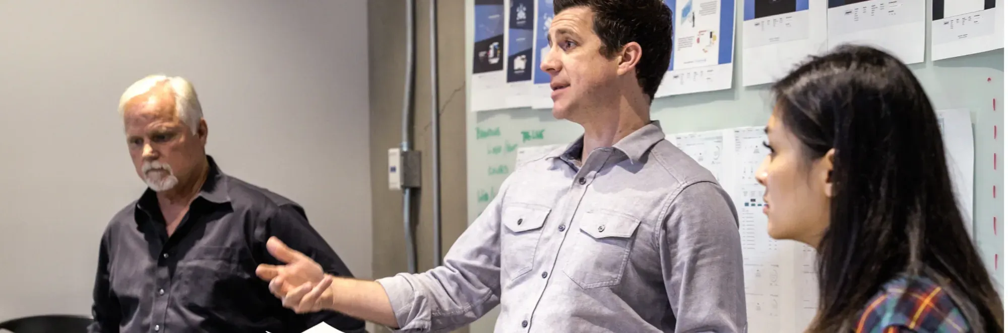 Three people having a discussion in a meeting room with charts and notes on the wall.