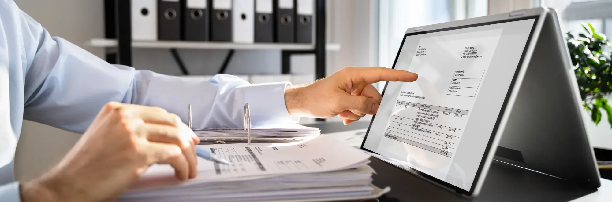 Person pointing at an invoice displayed on a tablet while reviewing printed documents in an office setting.