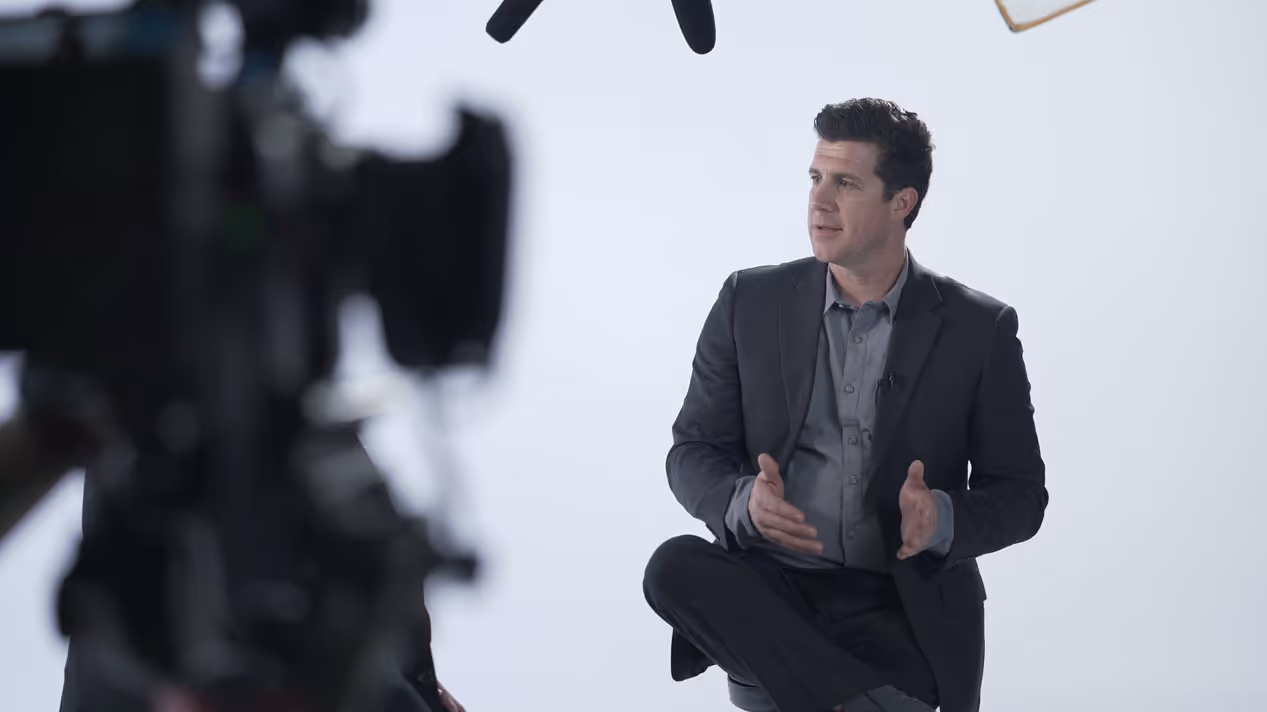 Man in a dark suit and gray shirt sitting and gesturing during an interview with a camera in the foreground.