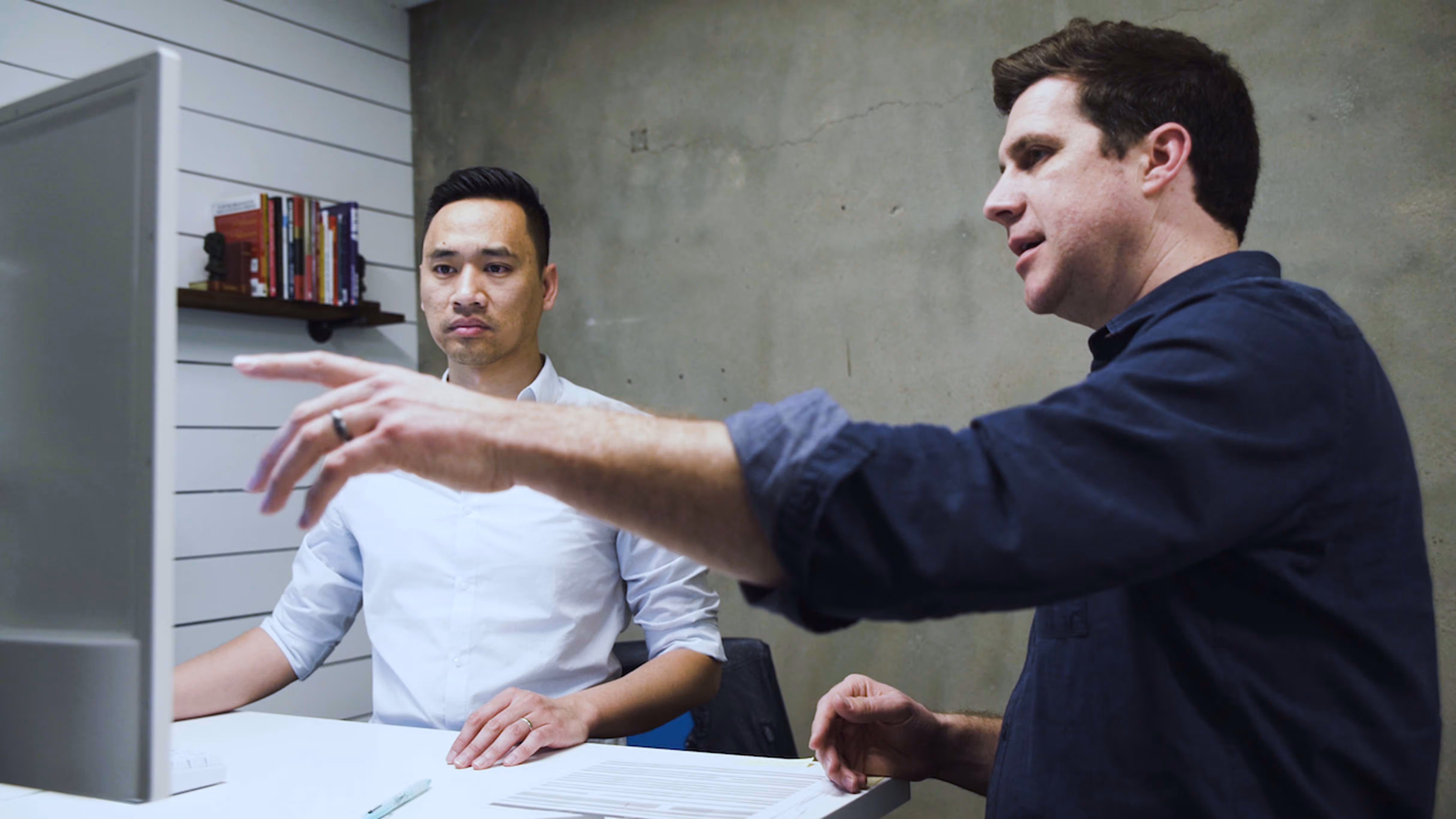 Two men sitting at a white desk looking at a computer monitor, one pointing at the screen while the other watches attentively.