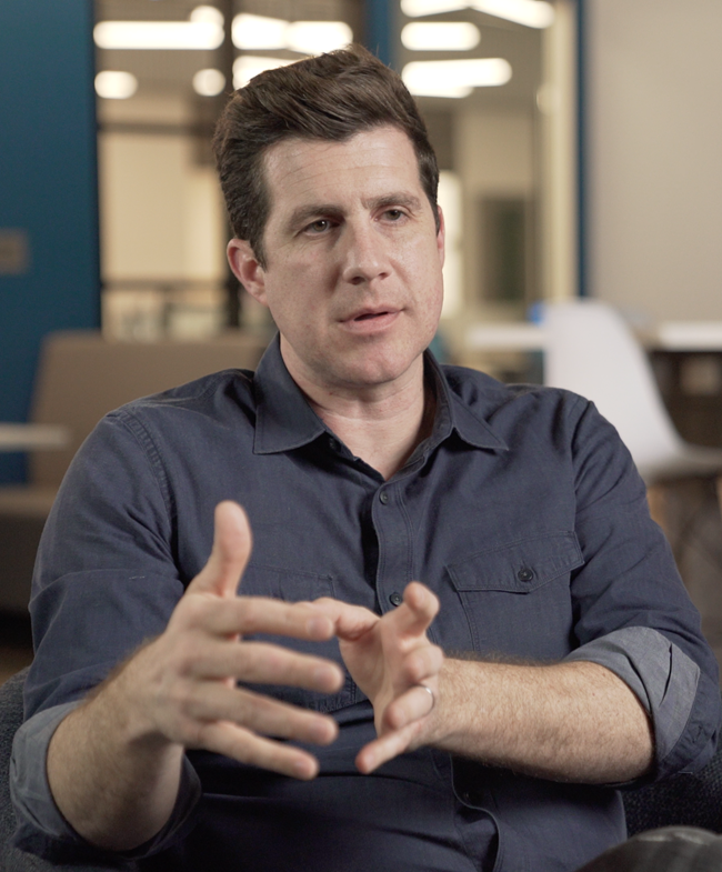 Man with short brown hair wearing a dark blue button-up shirt, gesturing with his hands while speaking in an office setting.