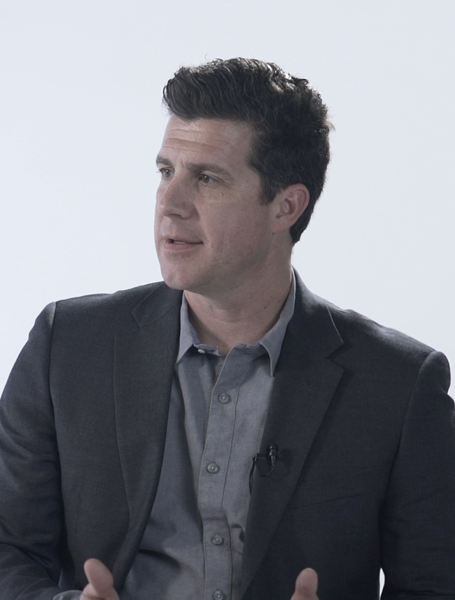 Man with dark hair wearing a gray button-up shirt and dark blazer looking to his left.