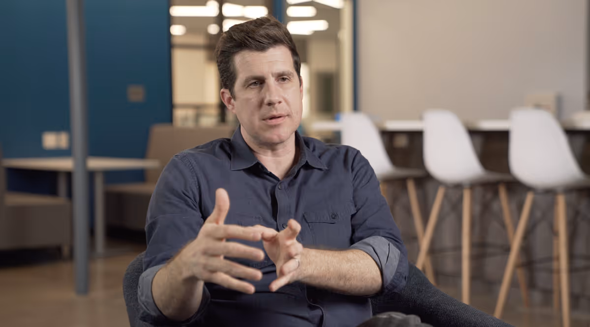 Man in a dark blue shirt sitting in a chair, gesturing with hands during a conversation in a modern office setting.